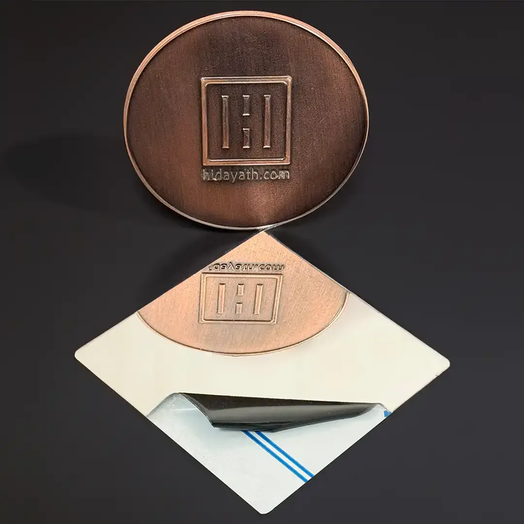 Plain mirror finish stainless swatch, diamond shaped, protective film peeled back at one corner, with an embossed Hidayath sample disc standing on it